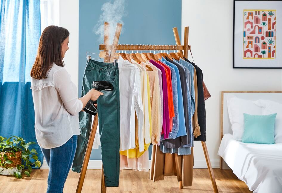Woman steaming clothes on a rack. A bed is in the background. Clothes of different colors.