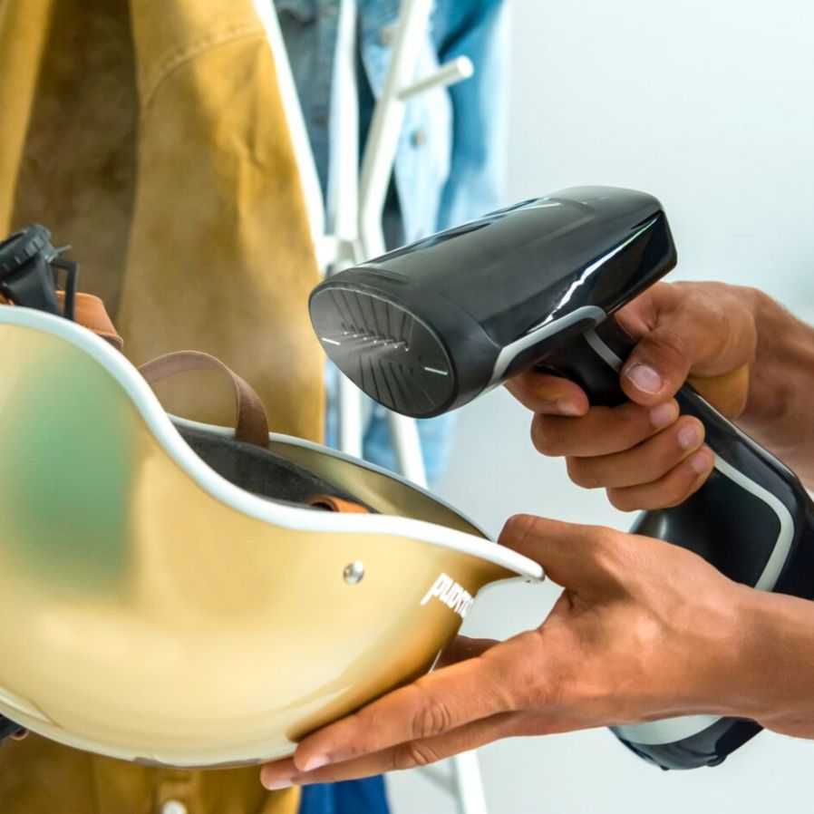 A person steaming a helmet with a handheld steamer. Background shows clothing.