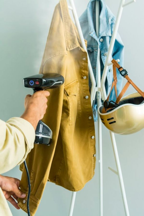 A person steaming a mustard-colored jacket. A denim jacket and helmet hang on a white rack.