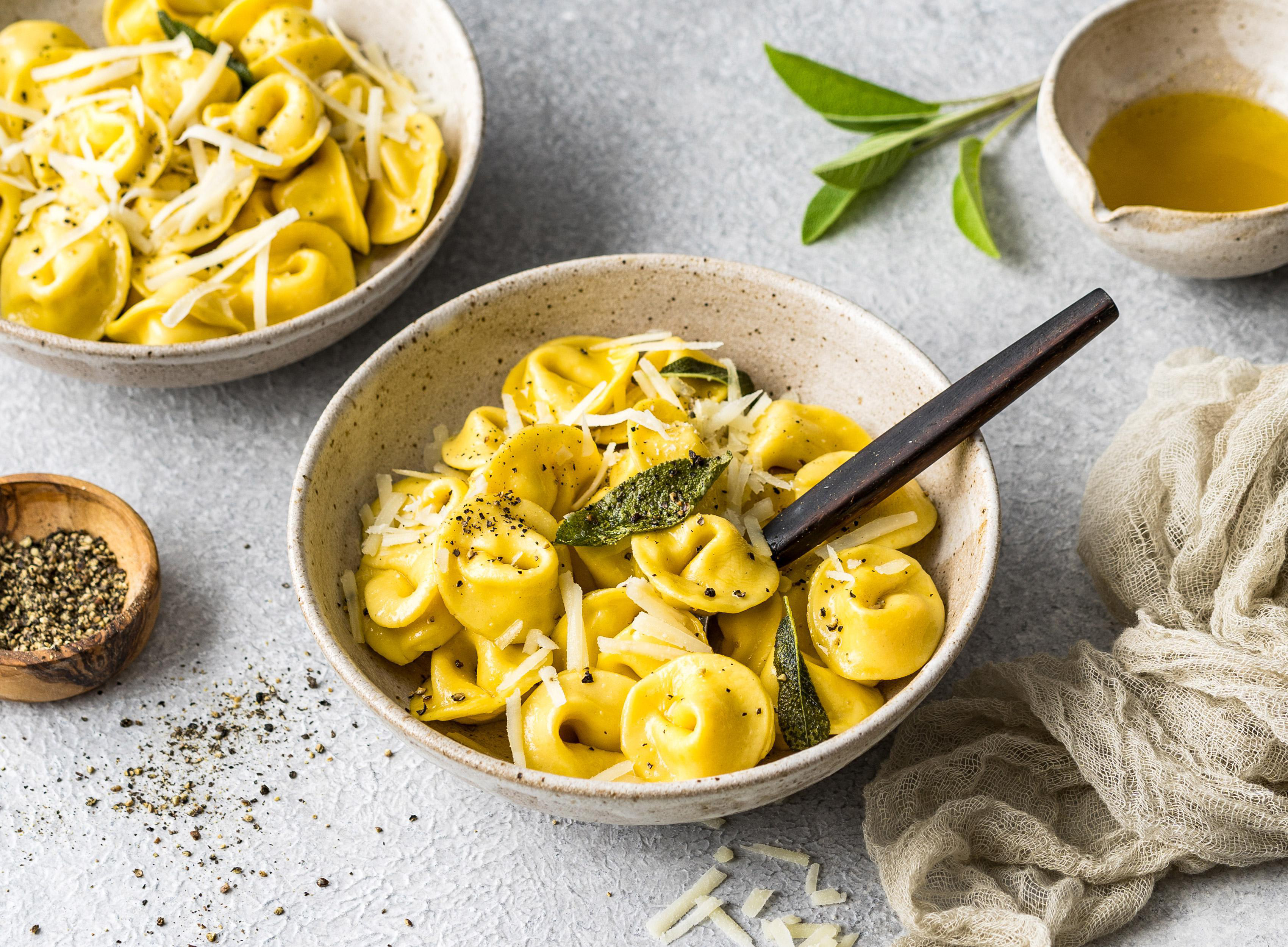 Zwei Schalen Tortellini-Nudeln mit Käse und Kräutern. Olivenöl und Pfeffer sind ebenfalls auf dem Tisch vorhanden.