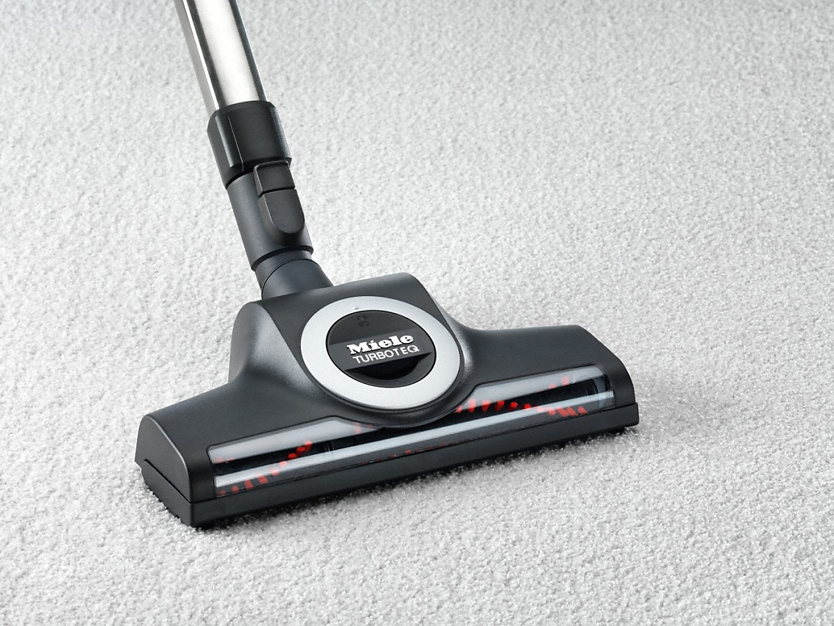 Vacuum cleaner head on a light gray carpet. The brush and Miele logo are visible.