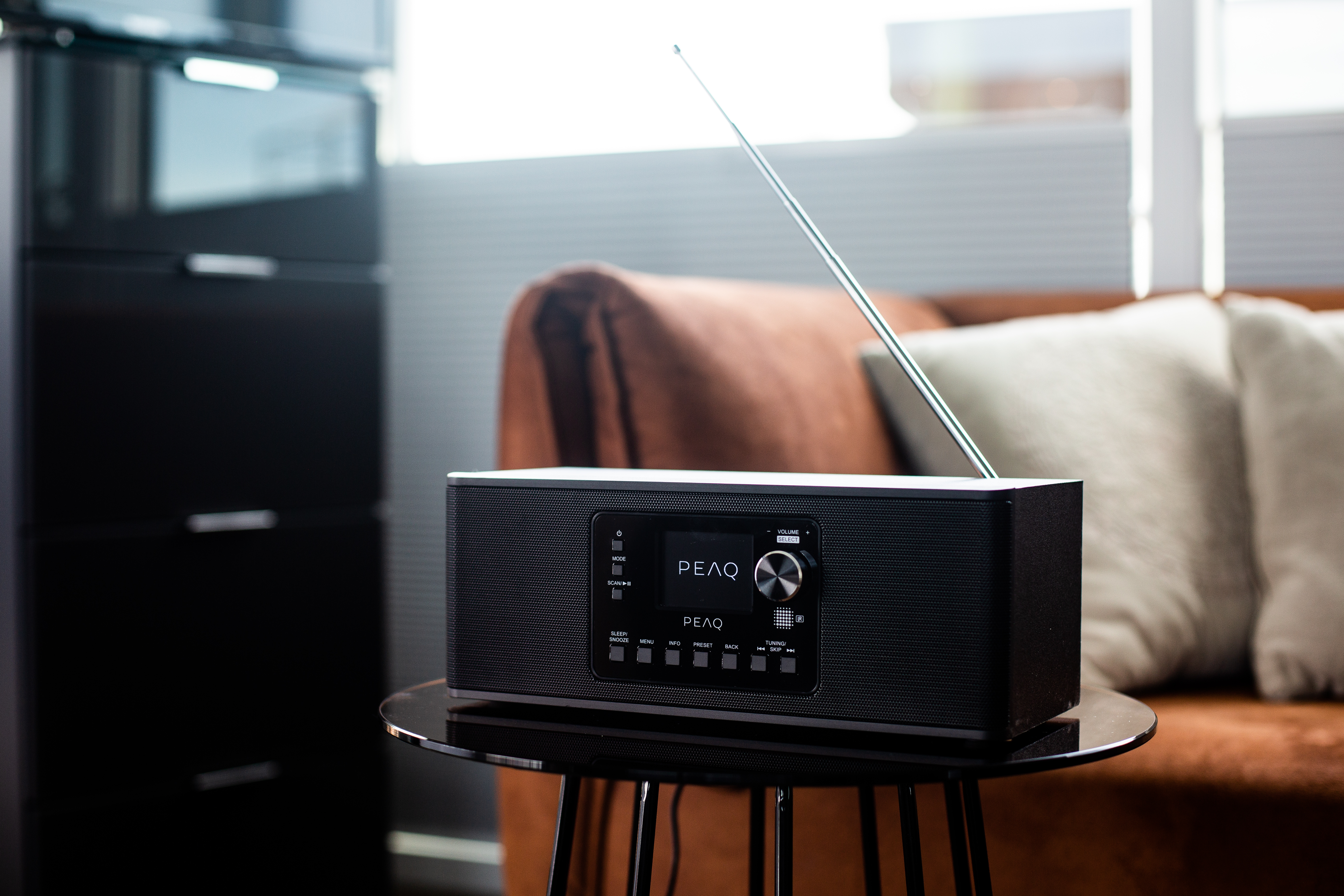 Schwarzes Radio auf einem Tisch. Es gibt einen Bildschirm und einen Drehregler. Im Hintergrund ein Fenster und Sofa.