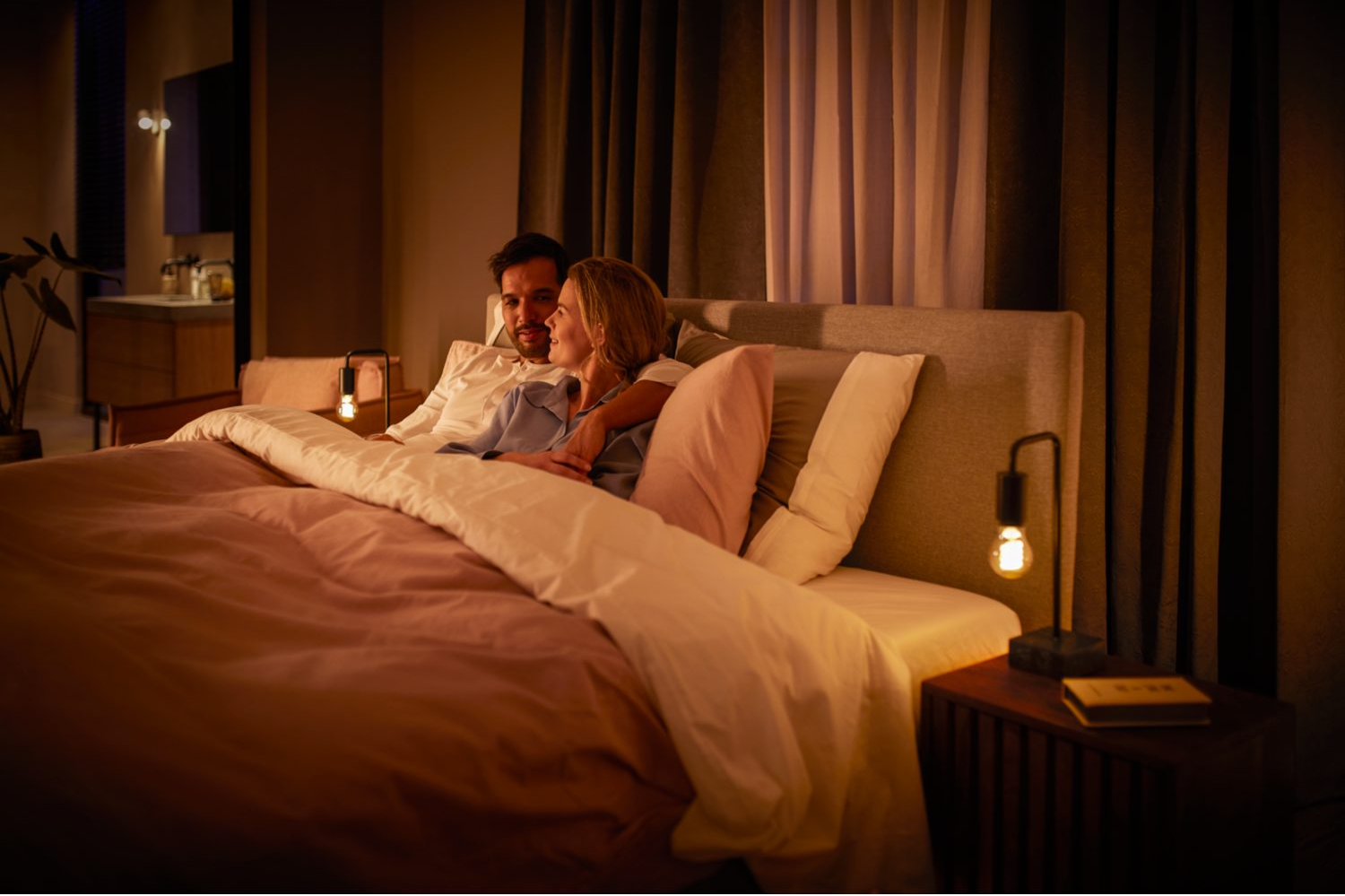 Un couple au lit avec des lampes de chevet allumées. Ils s'embrassent, une scène intime et chaleureuse.