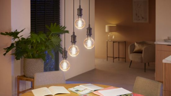 Scène intérieure avec lampes suspendues. Table ronde, plante et livres visibles. Éclairage chaleureux, tons neutres.