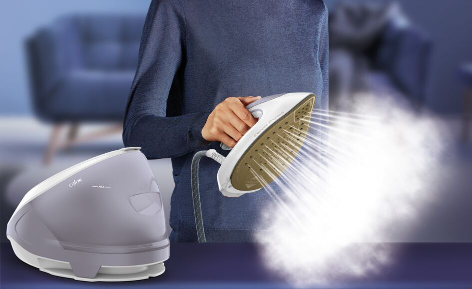 A woman holds a steam iron, steam emanating from it. A steam generator is on the left.