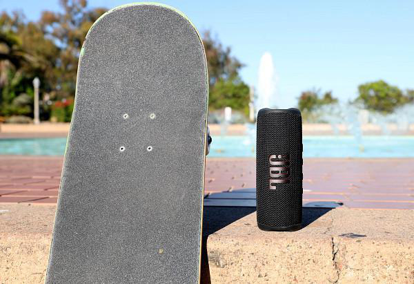 Skateboard und schwarzer Lautsprecher auf Stein, mit Brunnen und blauem Himmel.