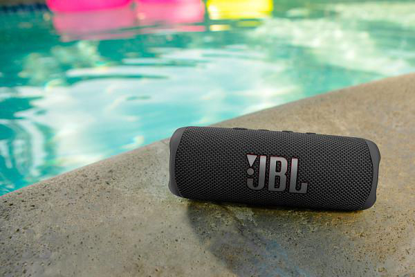 Ein schwarzer JBL-Lautsprecher liegt am Poolrand. Im Hintergrund befindet sich ein Pool mit rosa und gelben Schwimmkörpern.