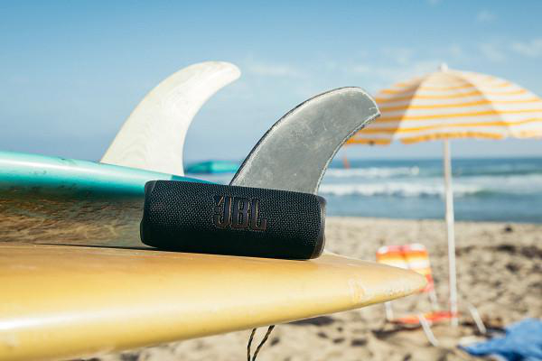 Ein schwarzer JBL-Lautsprecher liegt auf einem Surfbrett, flankiert von Flossen. Strandszene mit Sonnenschirm und Meer im Hintergrund.