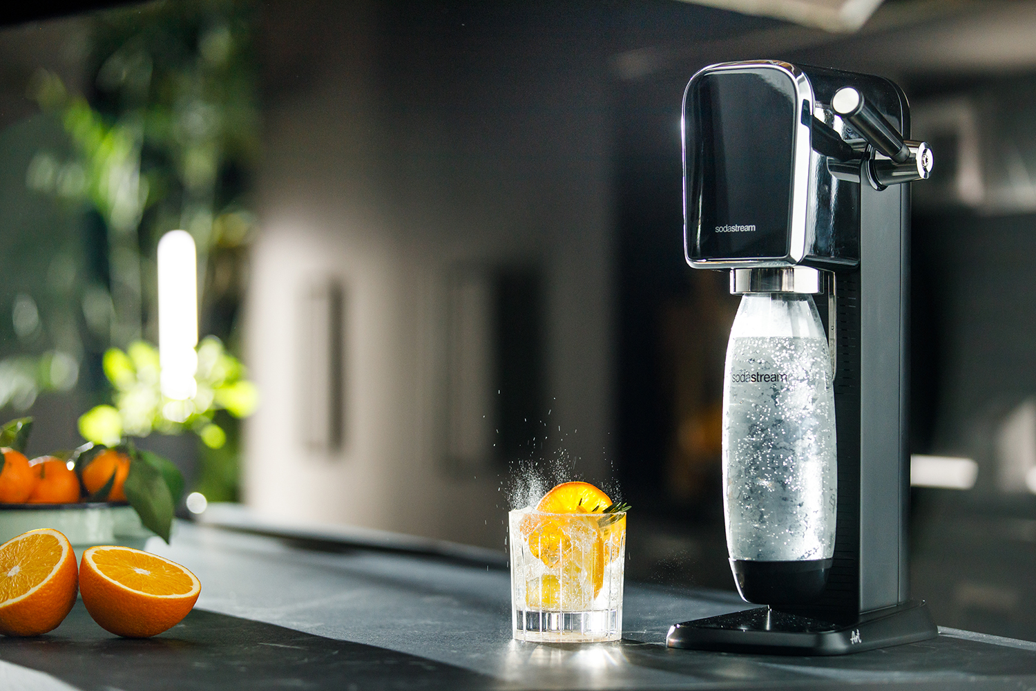 Een SodaStream machine met een glas water met sinaasappelschijfjes en ijs. Sinaasappels op tafel.