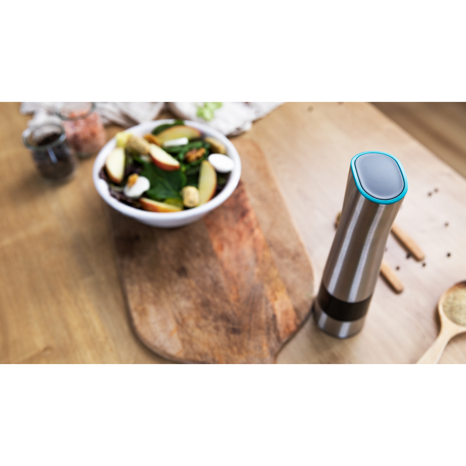 Un molinillo de acero inoxidable junto a una tabla de cortar de madera con un cuenco de ensalada.