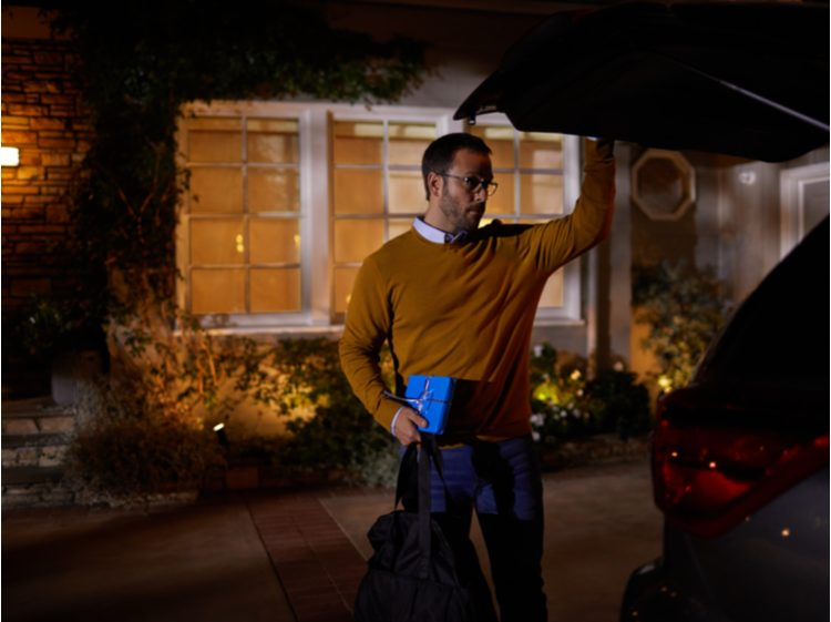 Homme chargeant le coffre d'une voiture la nuit. Il tient un cadeau et un sac. Maison avec fenêtres éclairées en arrière-plan.