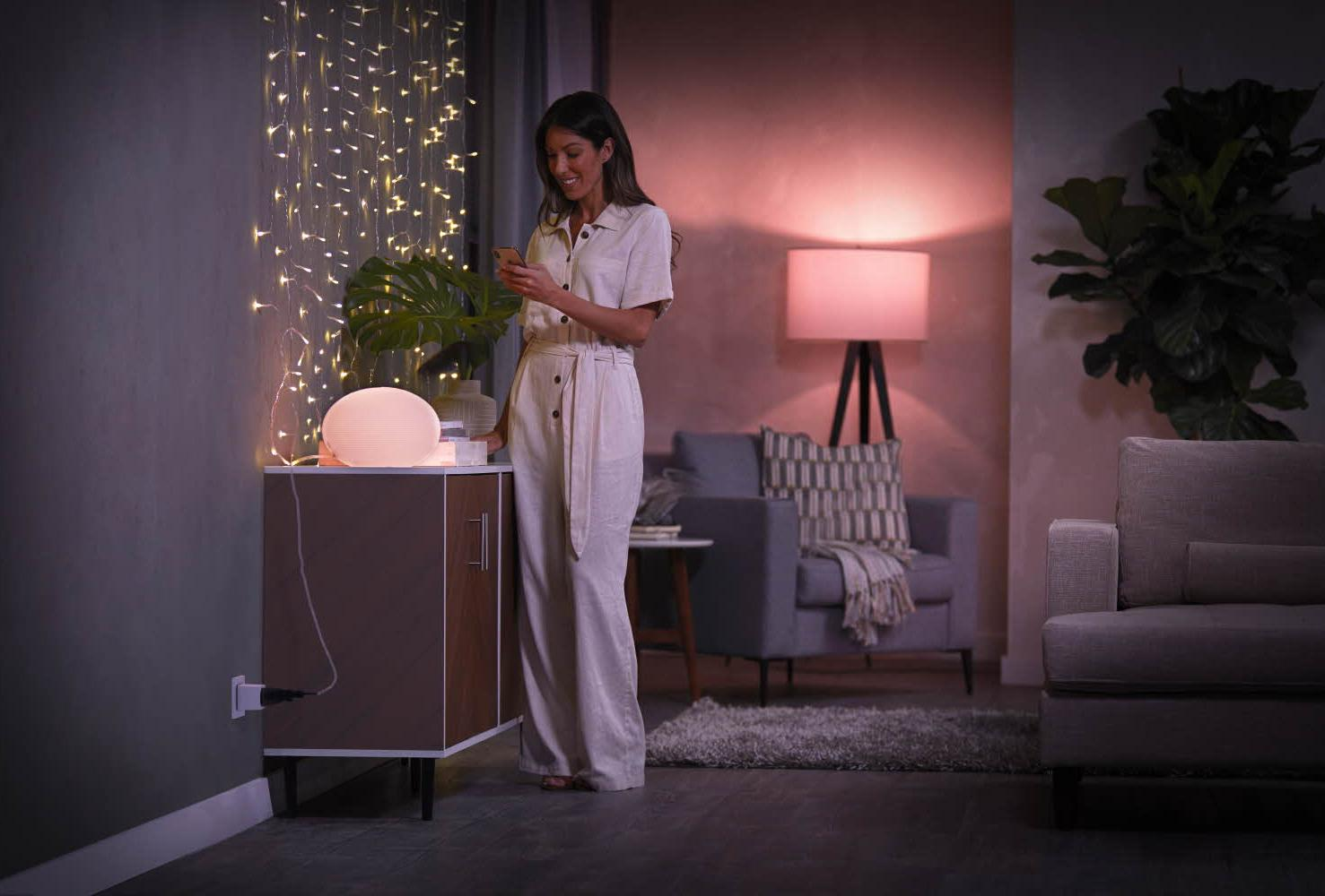 Femme dans une pièce avec des lumières et une lampe. Elle tient un téléphone près d'une table de chevet.