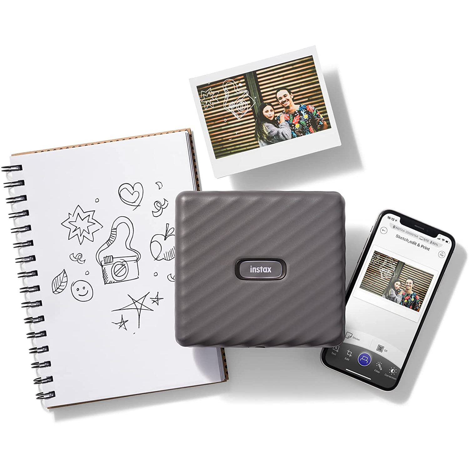 Una impresora Instax con fotos y un cuaderno con dibujos sobre una superficie blanca.