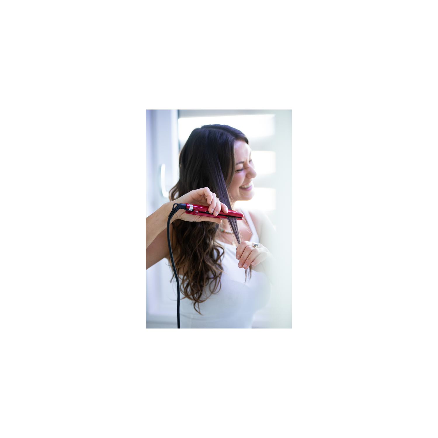 Mujer usando una plancha de pelo roja. Sonríe mientras se peina el cabello oscuro. Lleva un top blanco.