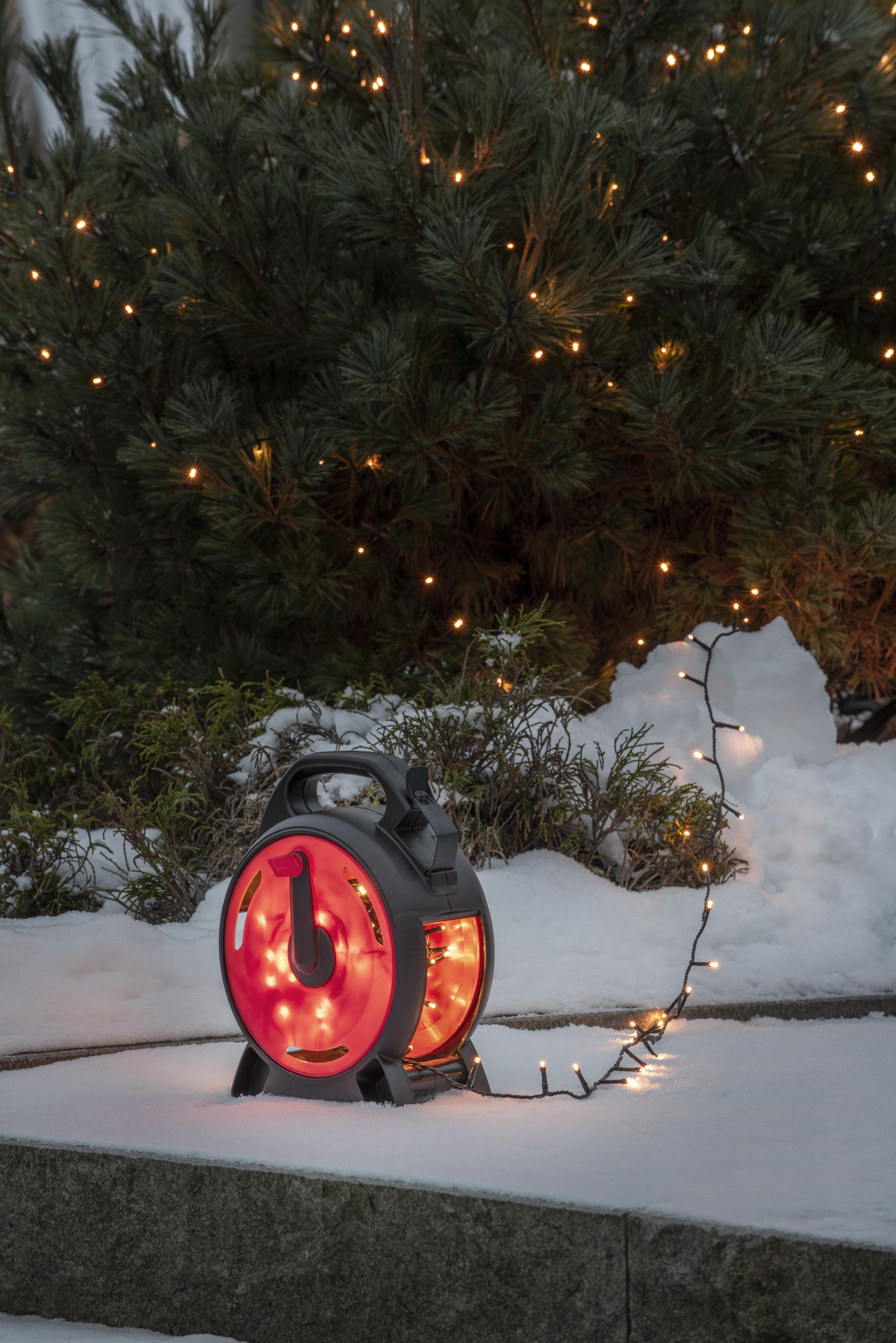 Eine rot-schwarze Lichtrolle leuchtet im Schnee. Eine Lichterkette erstreckt sich in Richtung eines Kiefernbaums.