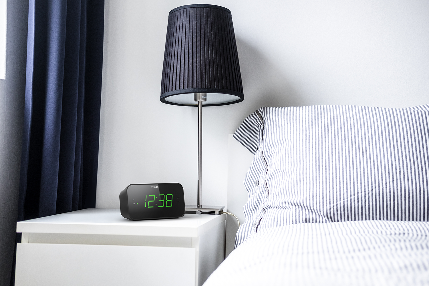 Black alarm clock on a white nightstand next to a bed with striped sheets.