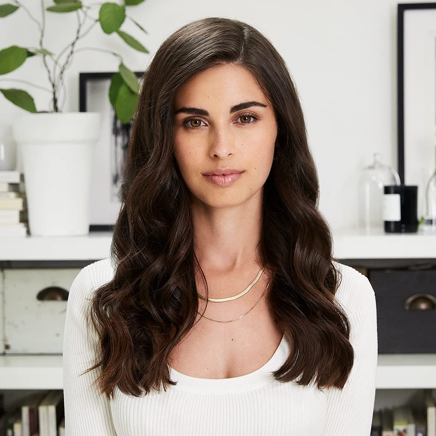 Una mujer con cabello largo y oscuro mira al espectador. Lleva una parte superior blanca y un collar dorado.