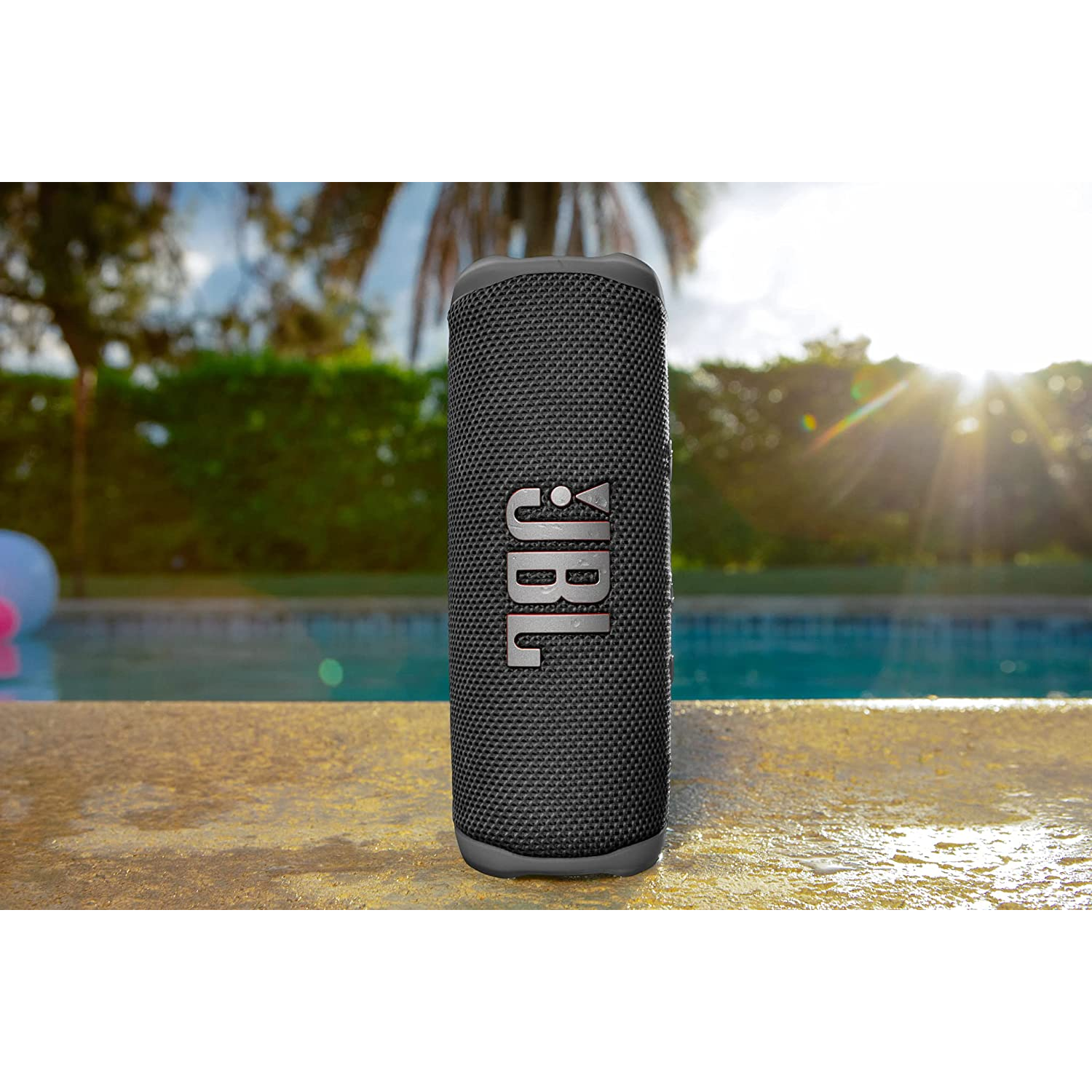 Altavoz JBL negro, vertical. De pie sobre una superficie de hormigón junto a una piscina con agua azul.