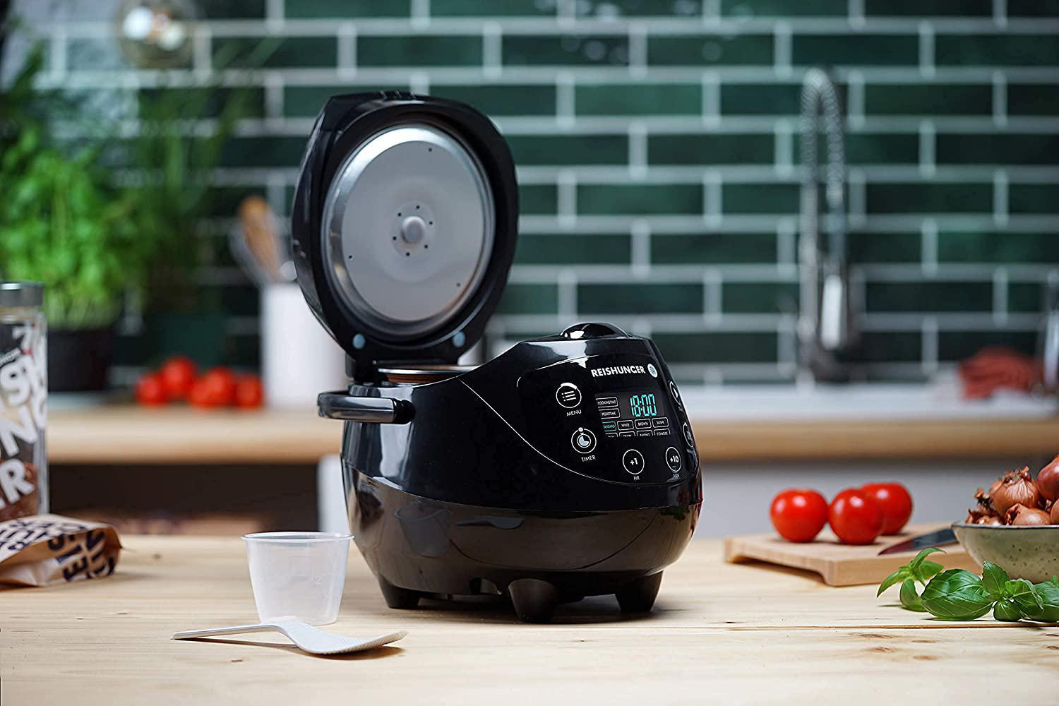 Schwarzer Reiskocher mit offenem Deckel. Daneben Tomaten und ein Spatel.