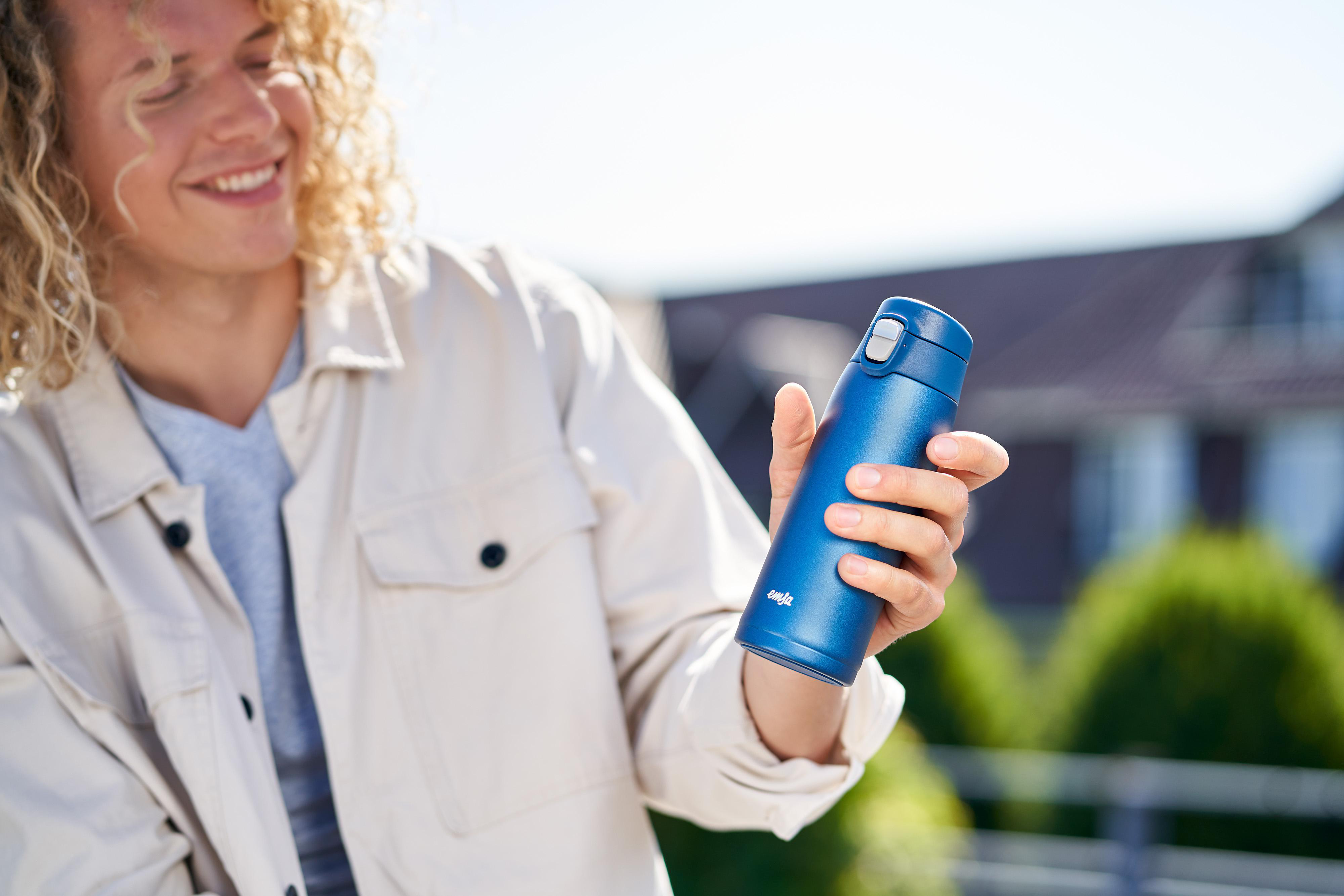 Ein Mann hält eine blaue Thermoskanne und lächelt. Hintergrund ist eine sonnige Szene im Freien.