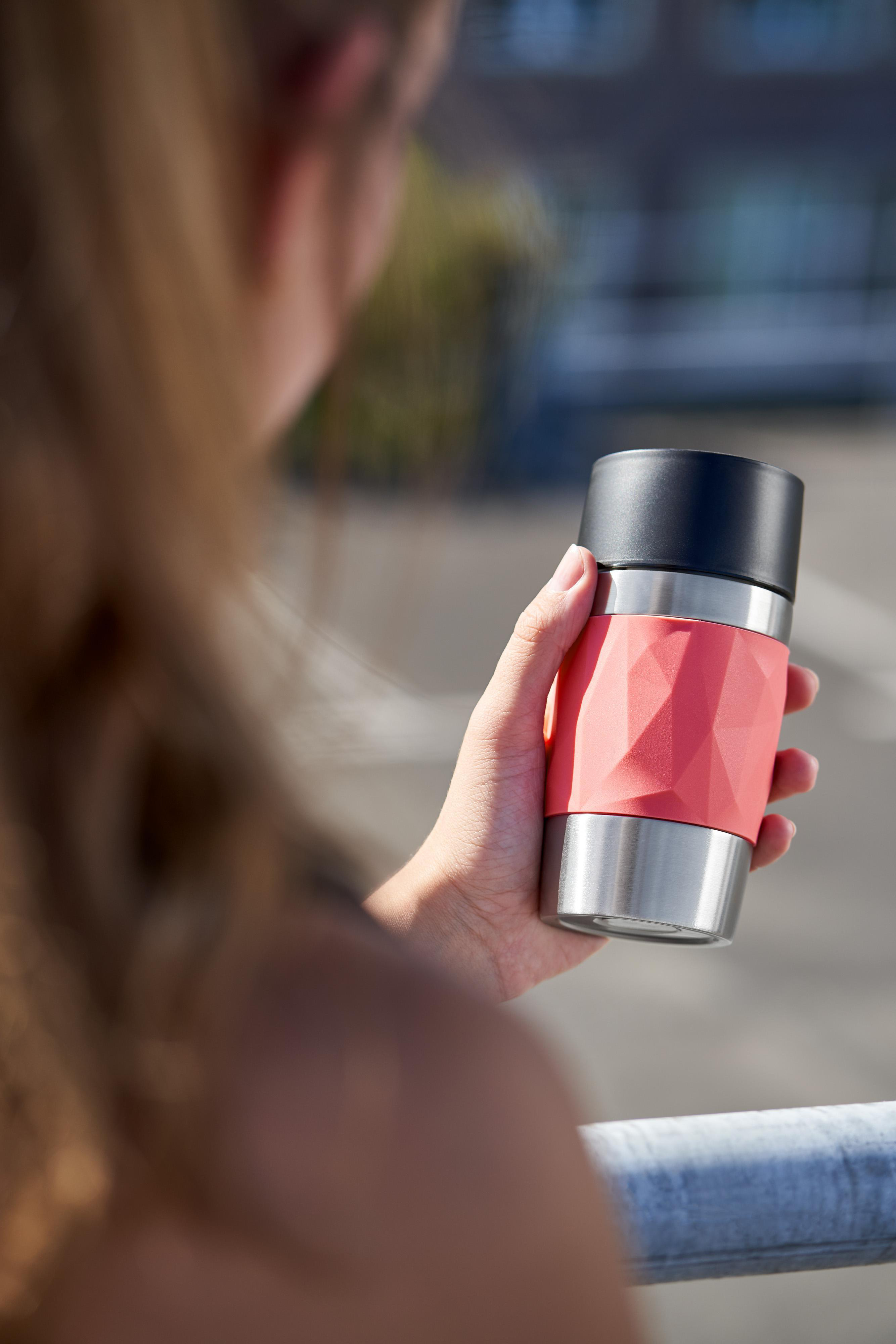 Frau hält einen Reisebecher mit rosa Abschnitt und silberfarbenem Boden, vor verschwommenem Hintergrund.