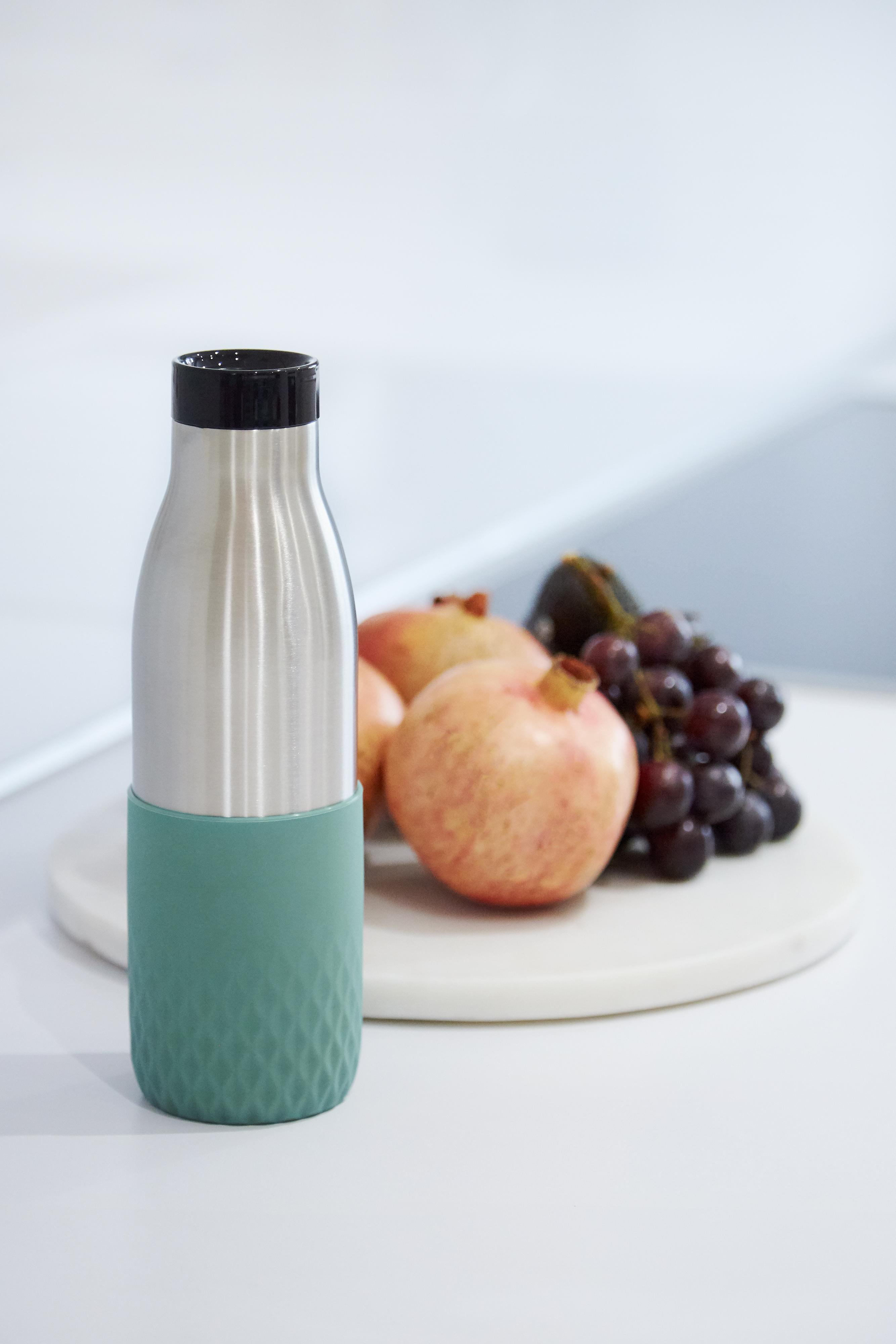Edelstahlflasche, grüner Sockel, mit Früchten auf Marmorbrett. Weißer Hintergrund.