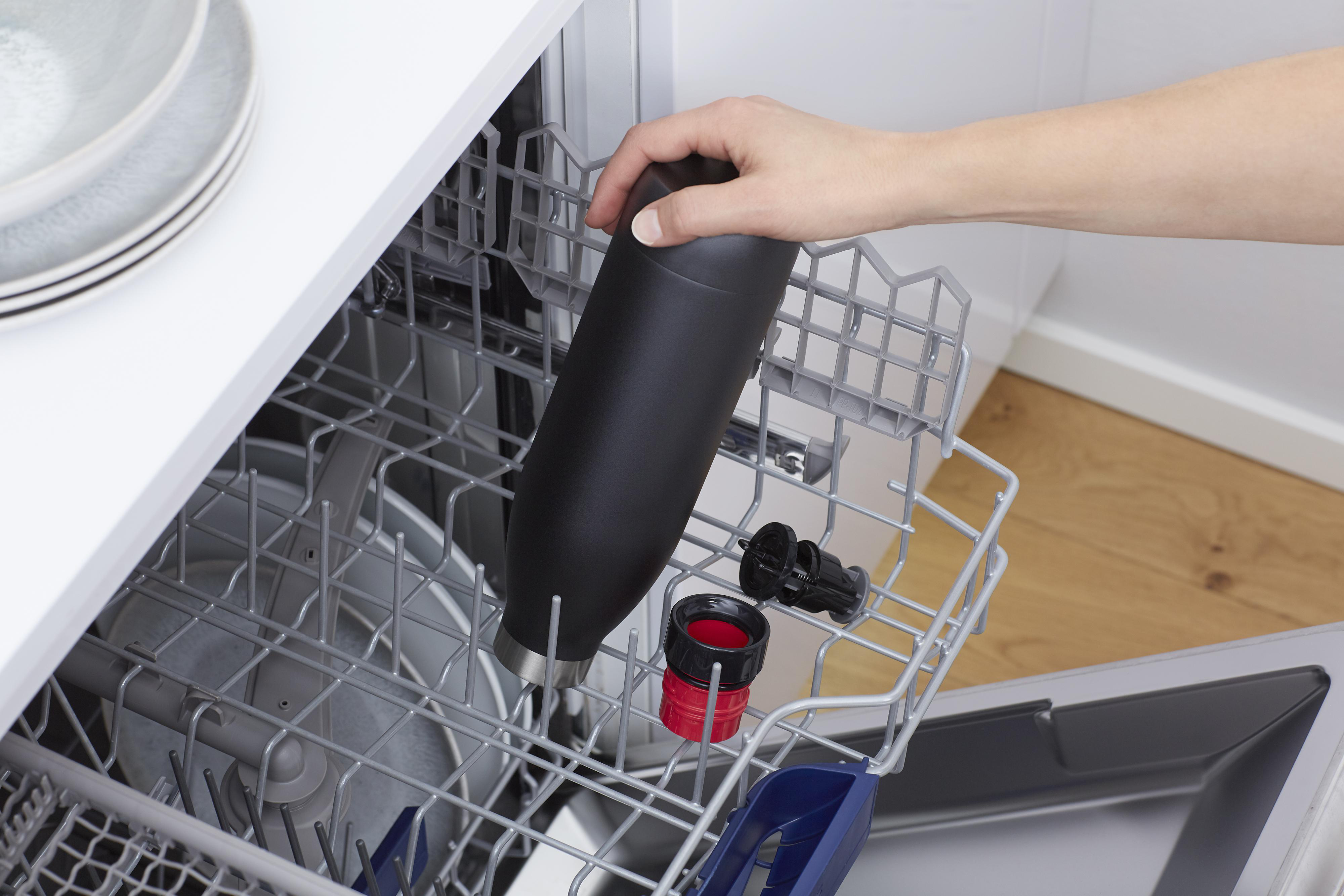 Hand legt eine schwarze Wasserflasche in einen Geschirrspüler. Teile werden in den Geschirrspüler gelegt.
