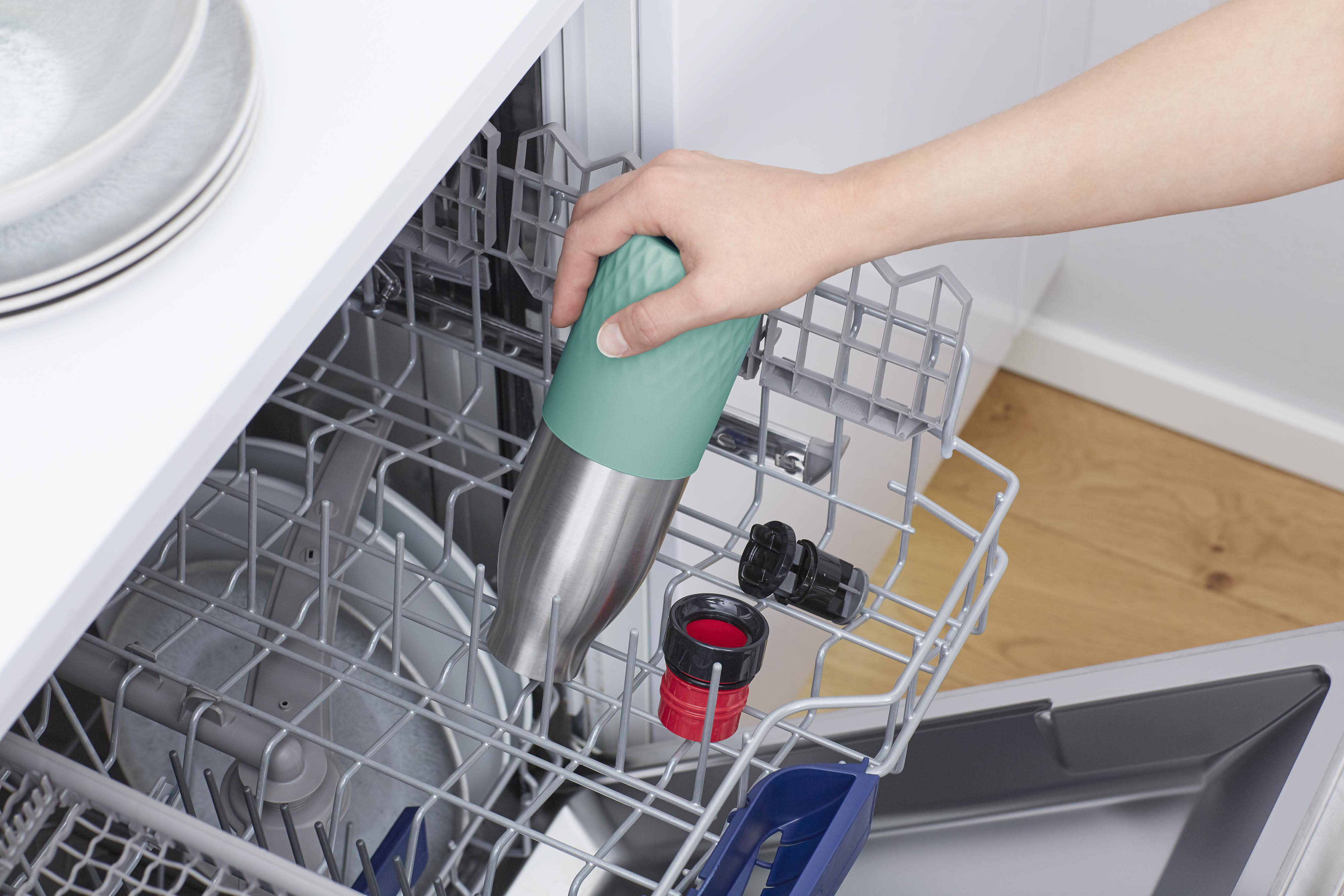 Hand legt eine grün-silberne Wasserflasche in einen Geschirrspüler. Andere Gegenstände befinden sich im Gestell.
