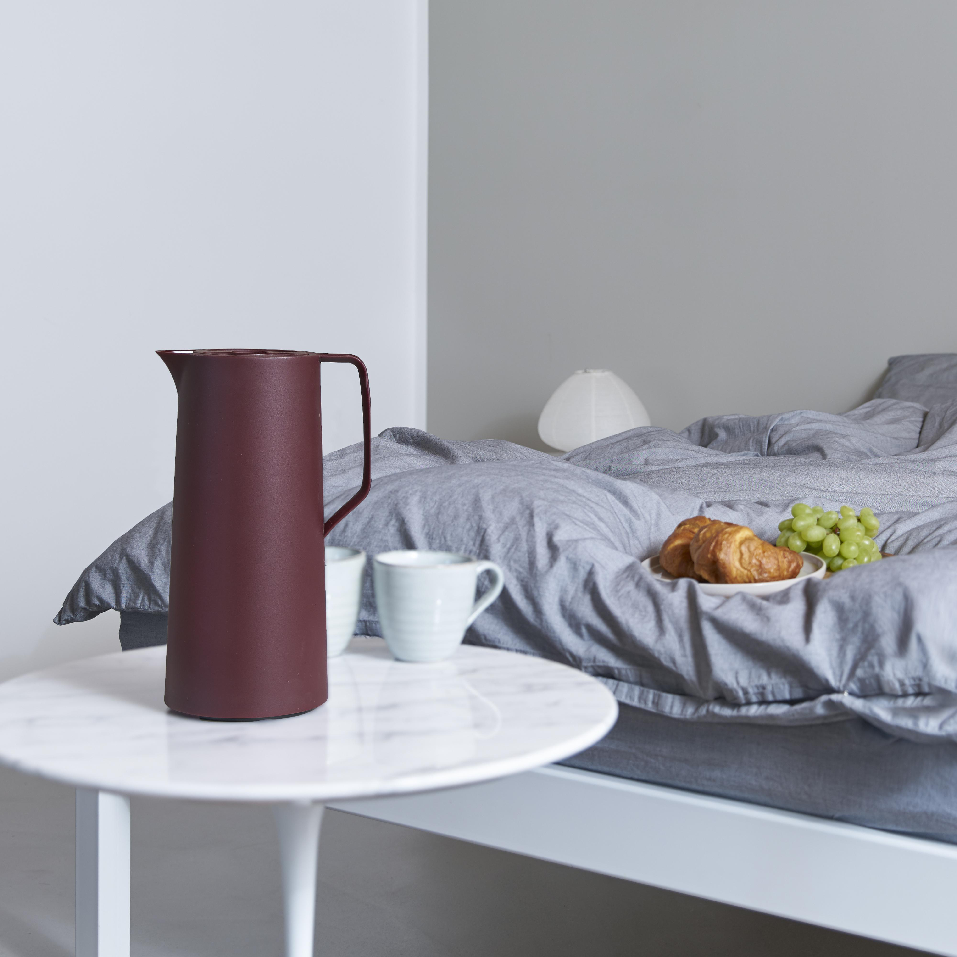 Weinrote Thermoskanne und zwei Tassen auf Tisch, Bett im Hintergrund.