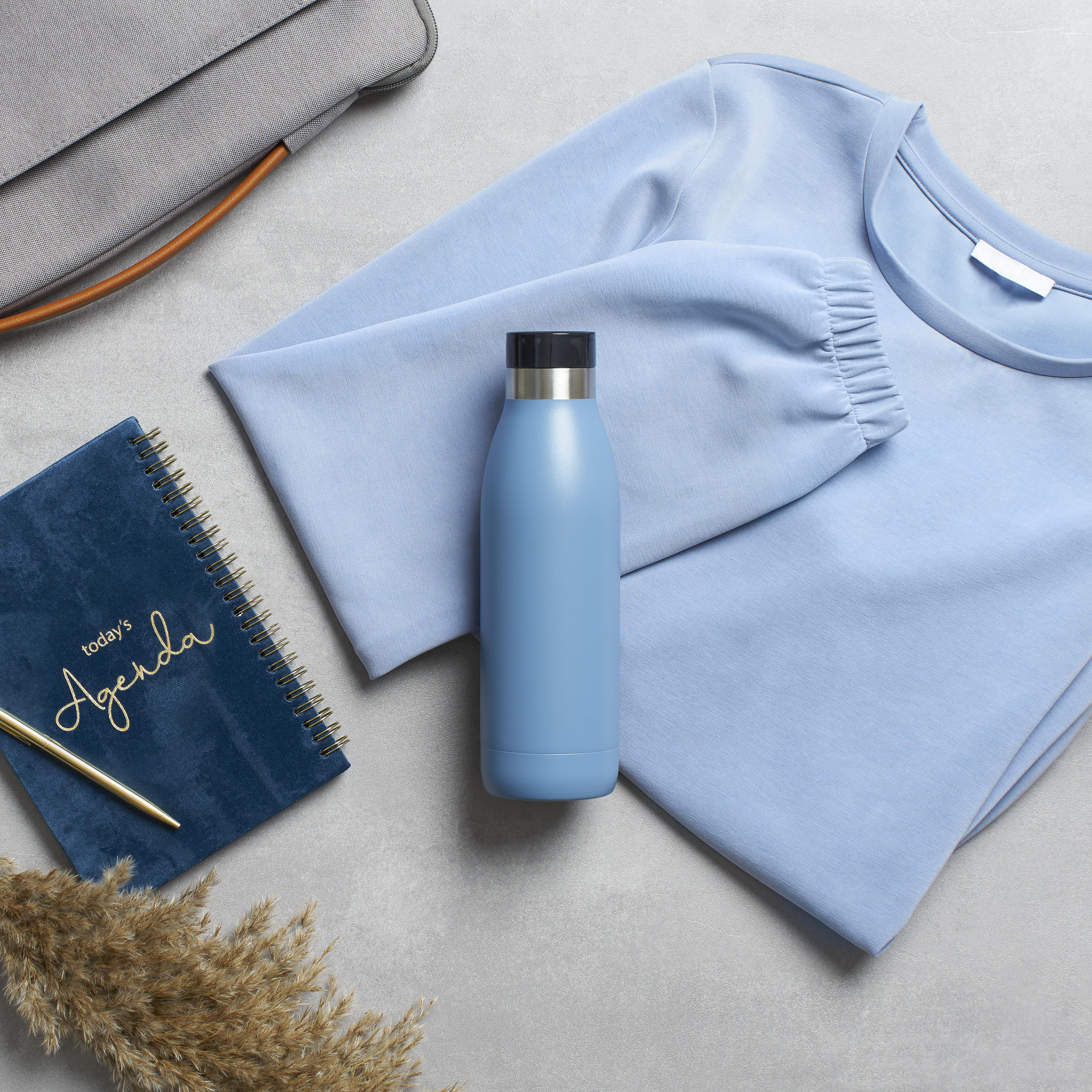 Blaue Wasserflasche, Notizbuch, Stift und Hemd auf grauer Oberfläche. Tasche im Hintergrund.