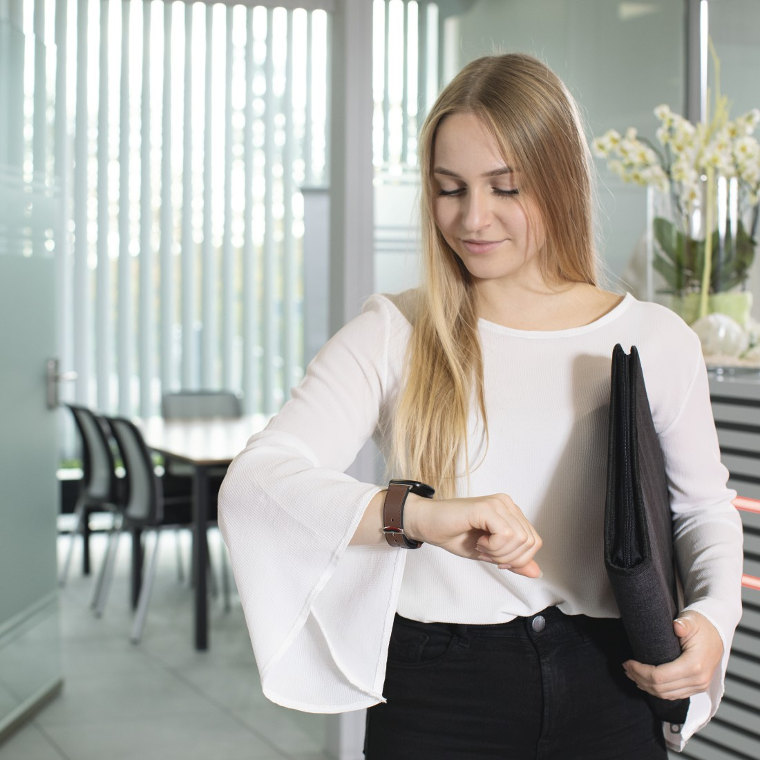 Eine blonde Frau in weißem Oberteil und schwarzer Hose betrachtet ihre Smartwatch in einer Büroumgebung.