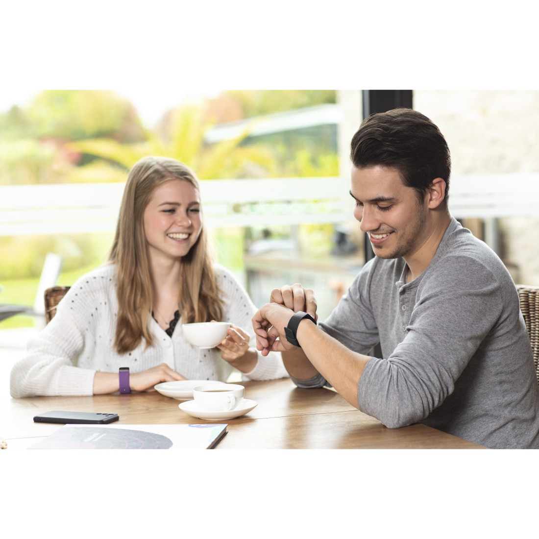 Ein Paar am Tisch, eine Frau hält eine Tasse, ein Mann schaut auf eine Smartwatch.