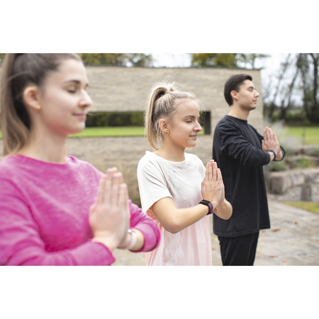 Drei Personen machen Yoga mit zusammengelegten Händen. Der Hintergrund ist ein Gebäude und grünes Gras.