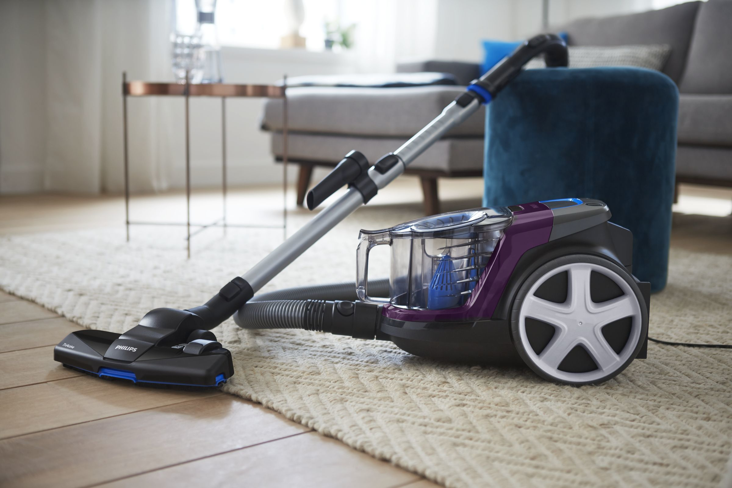 Purple and gray vacuum cleaner on a rug with a sofa in the background.