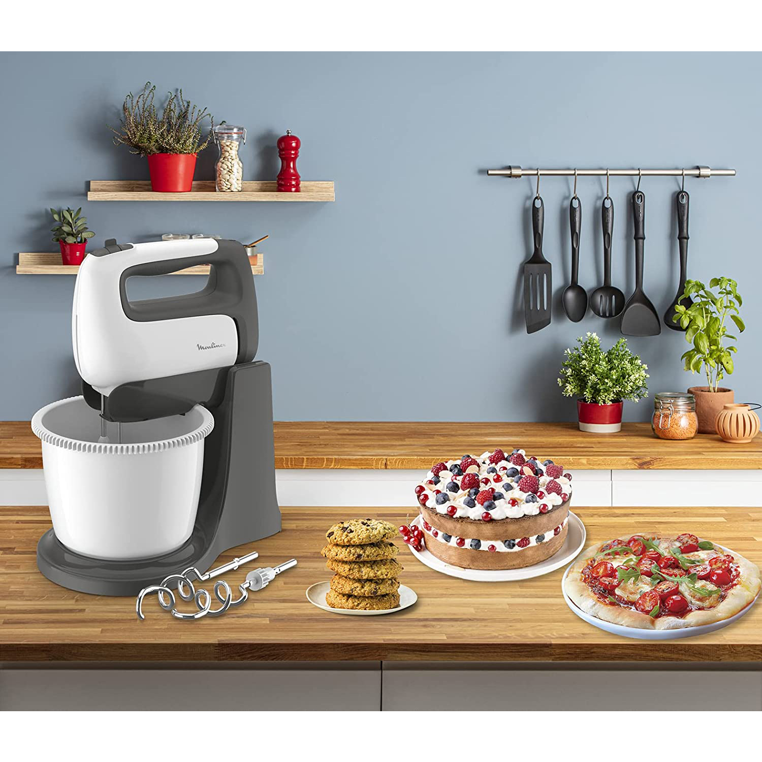 Scena in cucina. Un mixer bianco e grigio, biscotti, torta con frutti di bosco e una pizza con pomodori e basilico.