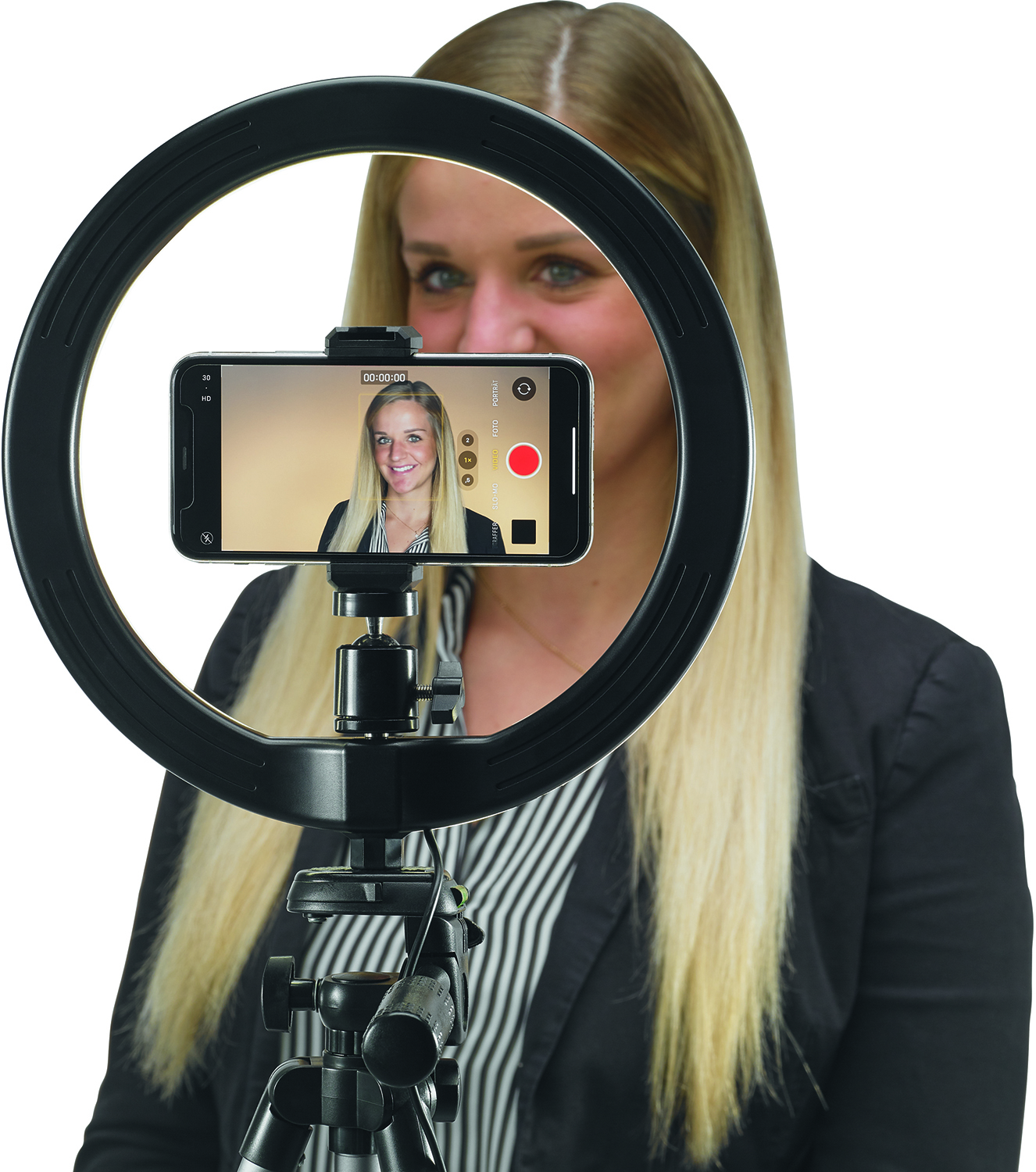 A woman filming herself with a phone on a ring light, smiling at the camera.