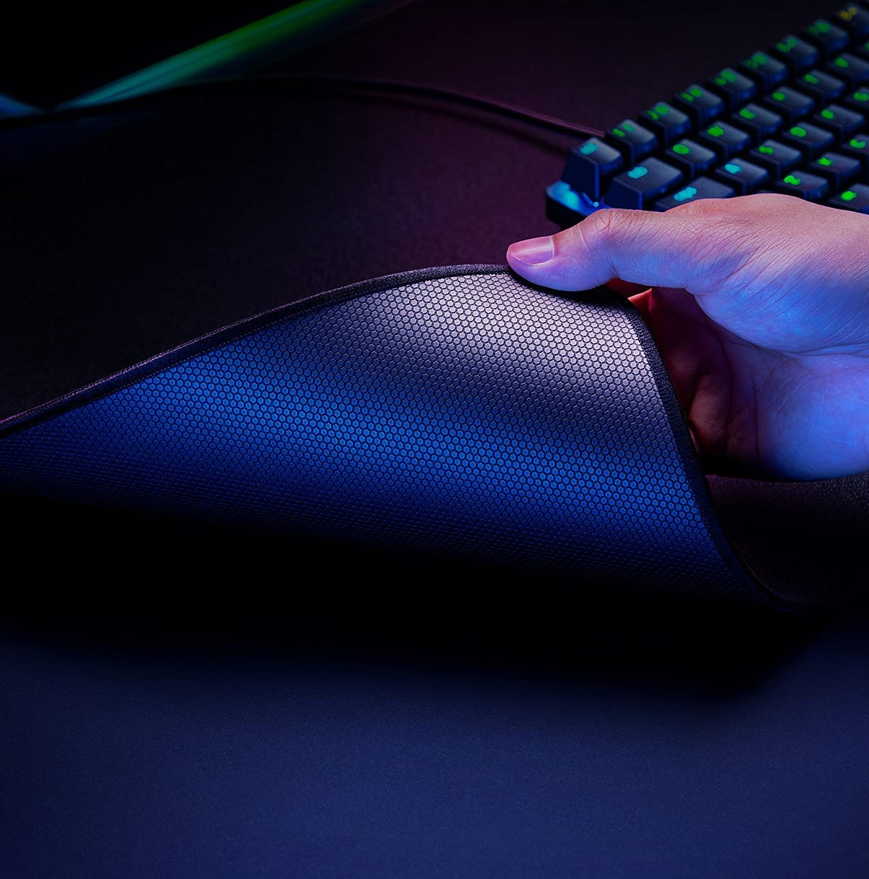 Une main soulève un tapis de souris gaming noir. Dessous avec motif en nid d'abeille, clavier en arrière-plan.
