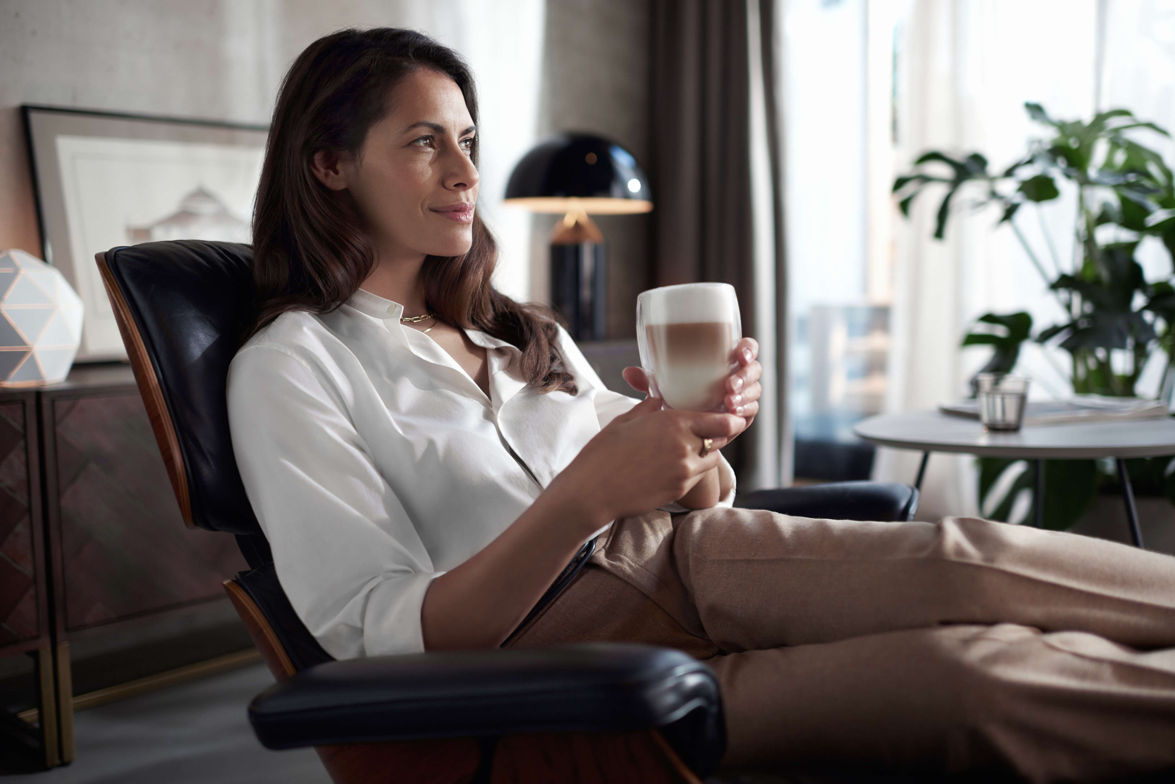 Donna su una sedia che tiene una tazza di caffè, guarda a sinistra, con una lampada e una finestra.