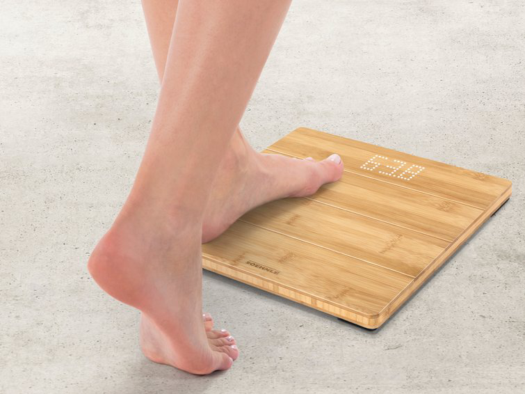 Pied de femme sur une balance en bois. Affiche 63.0 en blanc. Fond en béton.