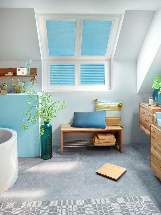 Une salle de bain avec un plafond en pente et une fenêtre avec des stores bleus. Un banc et des étagères en bois sont présents.