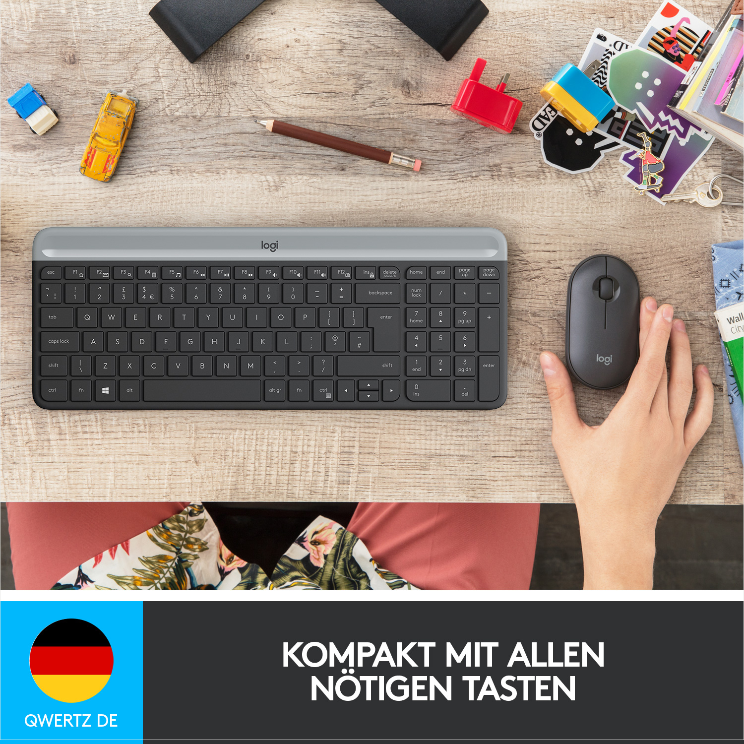 Eine graue Tastatur, eine Maus und kleine Spielzeuge auf einem Holztisch. Eine Hand schwebt über der Maus.
