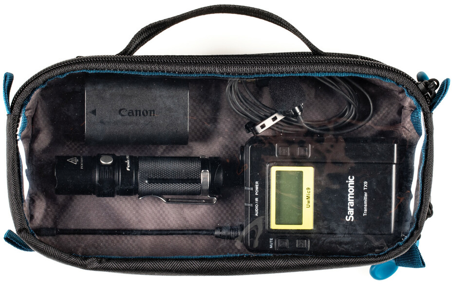 Eine transparente Tasche mit einer Canon-Batterie, einer Taschenlampe, einem Mikrofon und einem Saramonic-Sender.