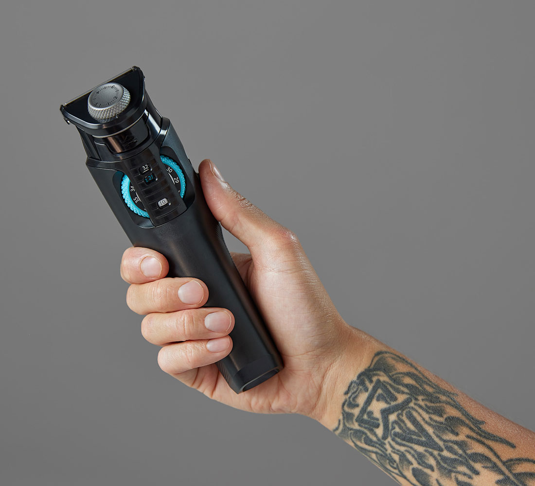 Hand hält schwarzen Barttrimmer. Der Trimmer hat ein blaues Rad und ein Tattoo auf dem Arm.