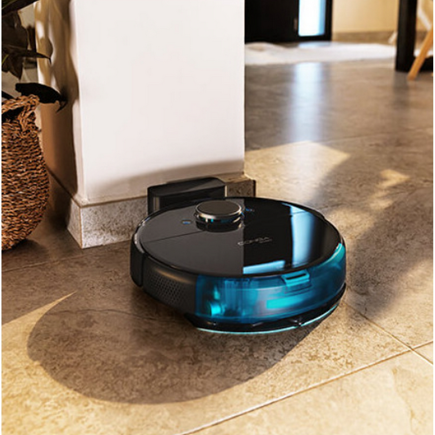 Aspiradora robot negra en suelo de baldosas, cerca de pared y cesta.