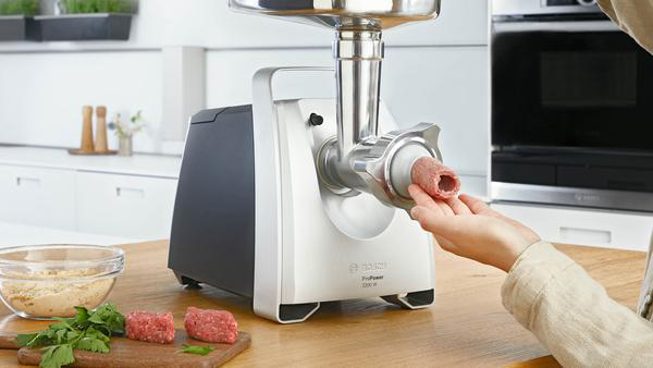 Ein Fleischwolf, mit einer Hand, die Fleisch in die Maschine führt, und Zutaten in der Nähe.