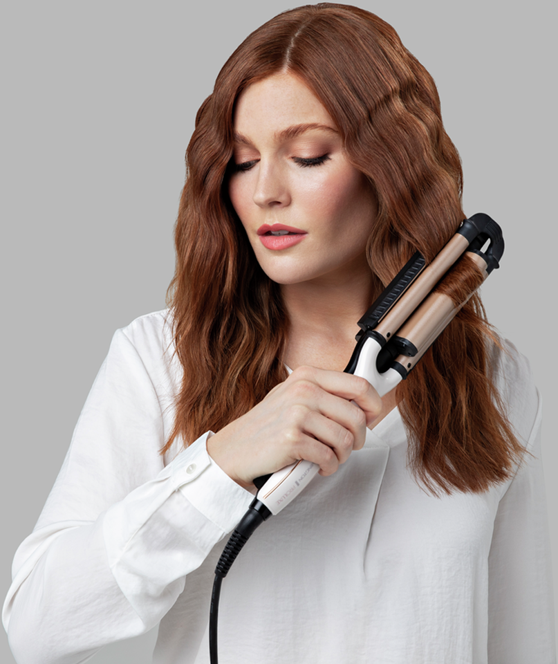 Femme aux cheveux roux utilisant un fer à onduler. Chemise blanche, fond gris.
