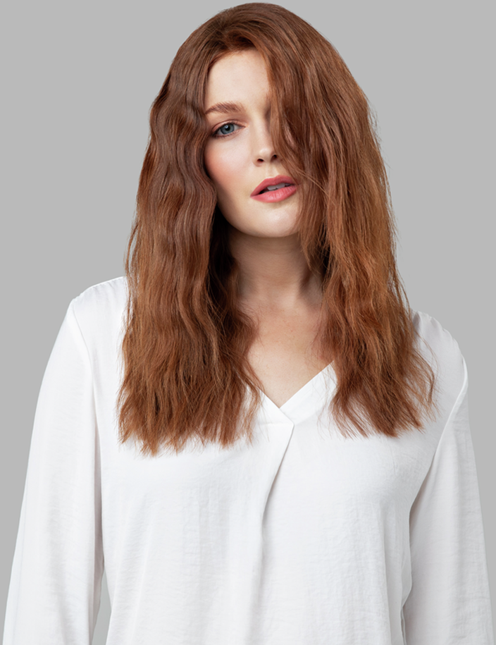 Une femme aux cheveux roux ondulés et une chemise blanche, regardant la caméra.