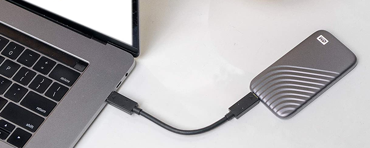 Egy laptop egy szürke hordozható merevlemezhez csatlakoztatva USB-kábellel, fehér felületen.