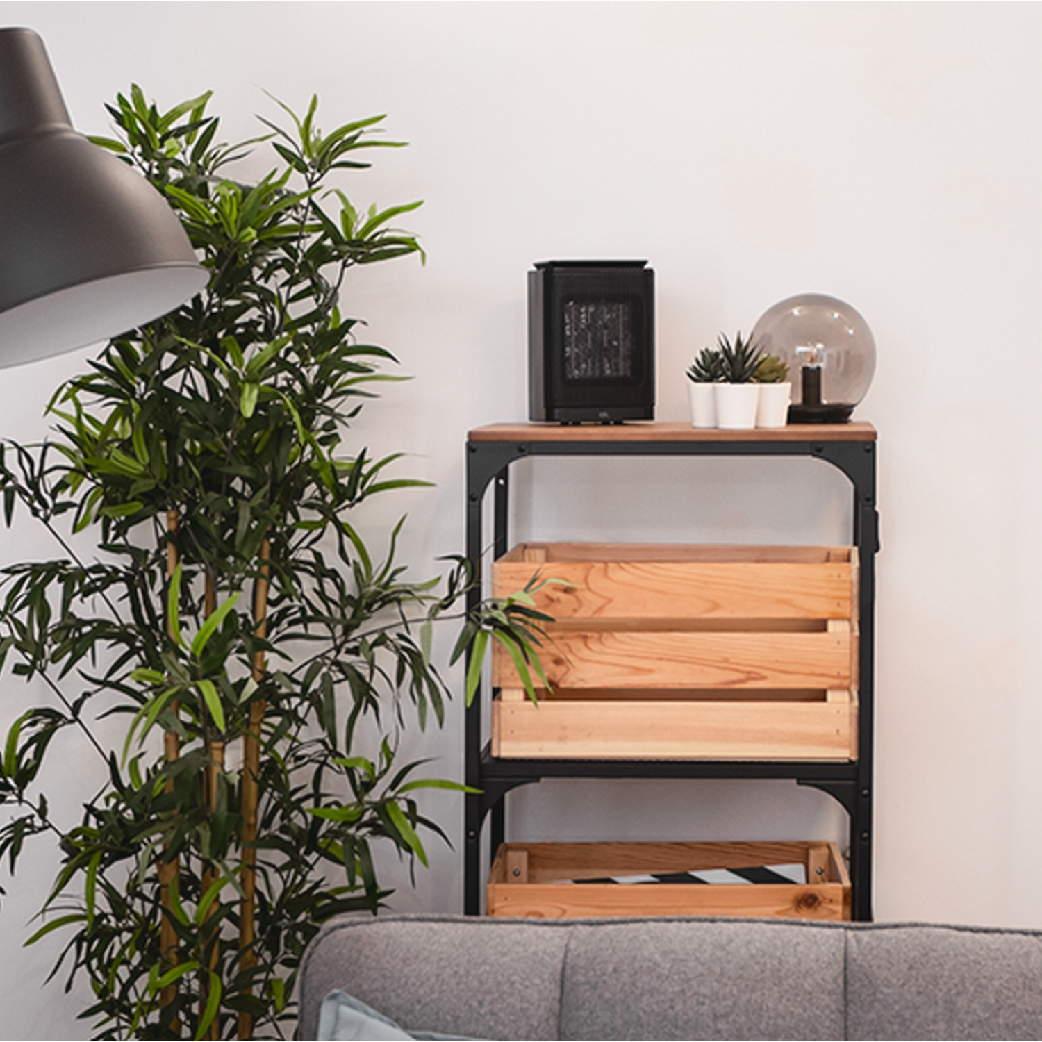 Un pequeño calefactor negro sobre una estantería de madera con plantas, una lámpara y un sofá al fondo.