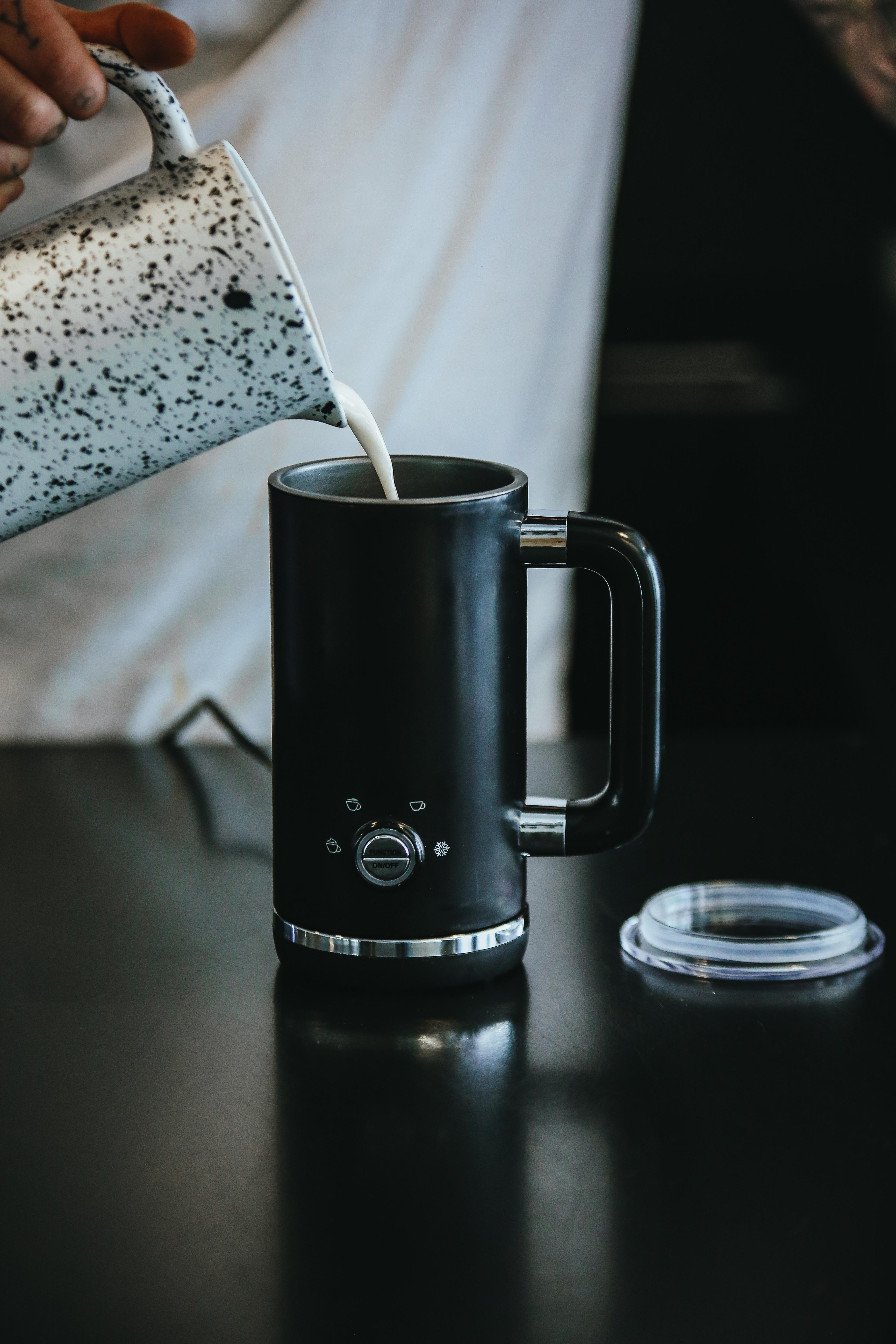 Eine Person gießt Milch aus einem gefleckten Krug in eine schwarze Kaffeemaschine.